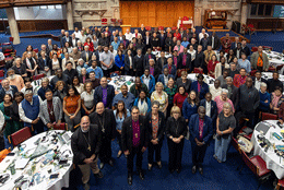 ACC delegates stand in a 19th century stone and wood-panelled guildhall in front of two ornate stained glass windows 
