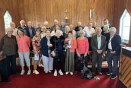 Around 30 people stand smiling in the wood-lined sanctuary of a country church flanked by two gothic peaked windows