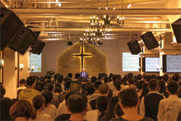 Industrial building filled with worshippers features pastor standing before large wall-mounted wooden cross