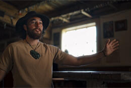 A Māori man with black hair and a hat with woven band speaks in a rural shed