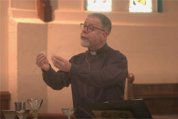 A grey-bearded Anglican priest holds up a consecrated wafer at the altar of a white-plastered church