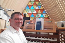 A dark-haired 50 year-old man in a white shirt sits at an organ console with a multicoloured triangular window in the background