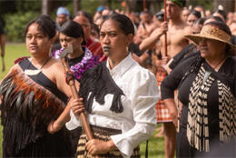 Three Māori women in challenge pose and kapa haka dress stand before their group on a sunny lawn