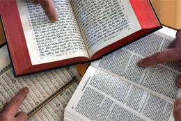 An open Torah, Bible and Quran with three fingers pointing to a passage of text in each