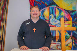 A male chaplain stands behind an altar with a dynamic painting of a dove and cross behind him
