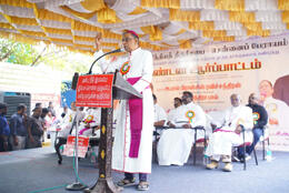 A South Indian Bishop stands at a microphone on an outdoor stage backed by a bench of Bishops and priests