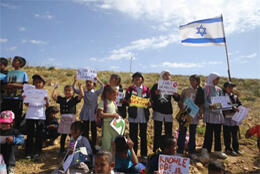 School children stand outside holding signs to allow them access to school, an israeli flags waves behind them
