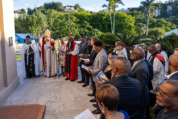 A group of Bermudan parishioners stand in a semi-circle before the doors of and historic church, with sunlit palm trees in the background 