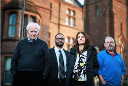 Three middle-aged men and one cwomen stand before a brick UK church