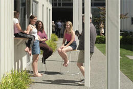 Three young women, a girl and an older man sit in the sun in a covered walkway next to a lawn