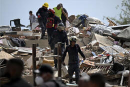 Emergency personnel in protective gear climb over rubble from a collapsed building