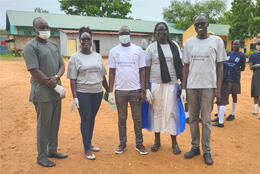 Five medical-masked and gloved South Sudanese Anglicans stand in a dirt courtyard wearing "Clear plastic pollution" t-shirts wearing "