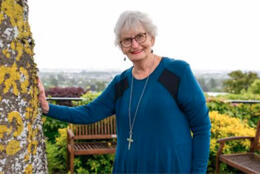 A woman with white hair and a blue tunic stands by a large tree in a garden