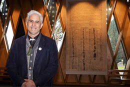 Māori Anglican male priest with white feather clerical collar stands beside a banner depicting the treaty of Waitangi