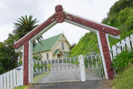 A cream coloured timber church Māori carved gateway arch with a green steel roof is seen through a  
