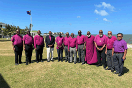 A dozen bishops in purple cassocks stand on a grassy field in the bright sun