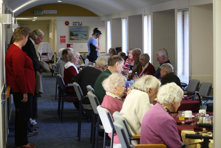Bishop Matthews says grace for the Churchill Courts residents, as they prepare to eat their evening meal in a corridor.
