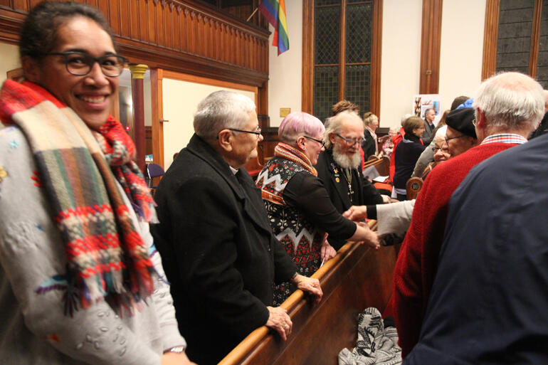 Rev Mele Prescott turns to the camera as her pew mates pass the peace.